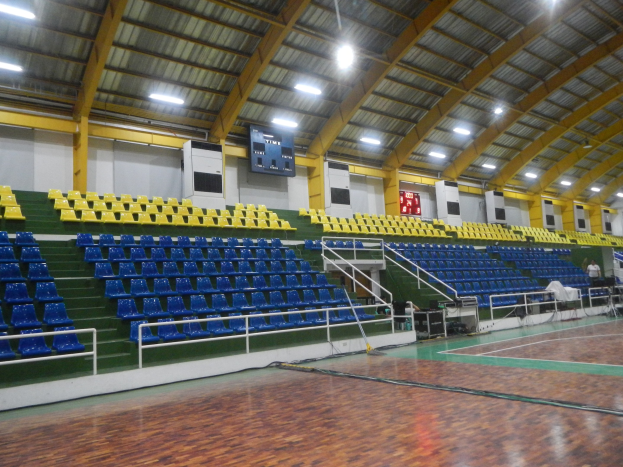 Große Sporthalle mit Holzboden, blauen und gelben Sitzplätzen, Geländern, Treppen mit Geländern, wandmontierten Bildschirmen, Deckenleuchten und einigen Menschen.