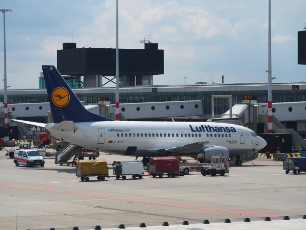 Lufthansa Airbus A320-200-Flugzeug auf dem Frankfurter Flughafen geparkt mit Bodenfahrzeugen und Wagen vorne, ein Gebäude und Laternenpfähle im Hintergrund unter einem klaren blauen Himmel.