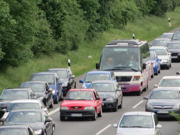 Stau auf einer Autobahn mit vielen Autos und einem Lieferwagen, Menschen in den Fahrzeugen, mit Bäumen und Gras im Hintergrund.