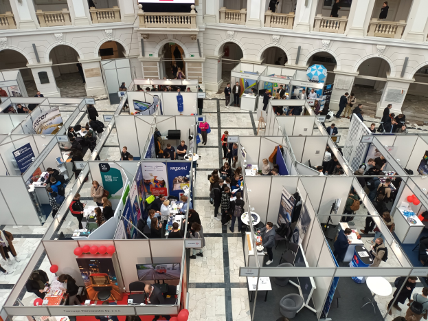Eine große Gruppe von Menschen auf einer internationalen Fachmesse mit Ständen, Bannern, Tischen, Stühlen, Luftballons und einem Gebäude mit Bögen, Säulen, Geländern und einem Hintergrundbild.