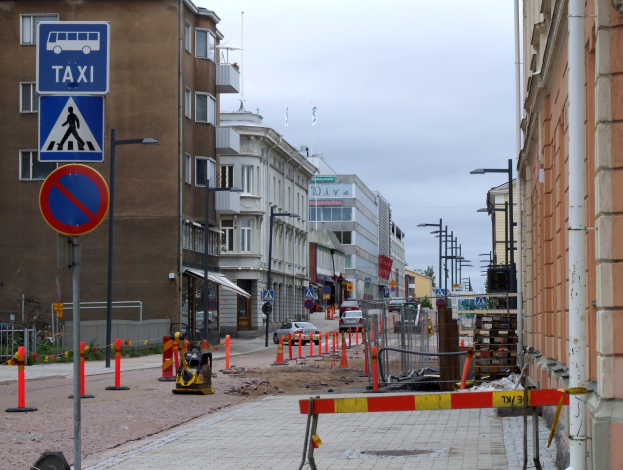 Stadtstraßenszene mit Gebäuden, Straßeninfrastruktur, Fahrzeugen, Bäumen und einer Baustelle mit Verkehrszeichen.