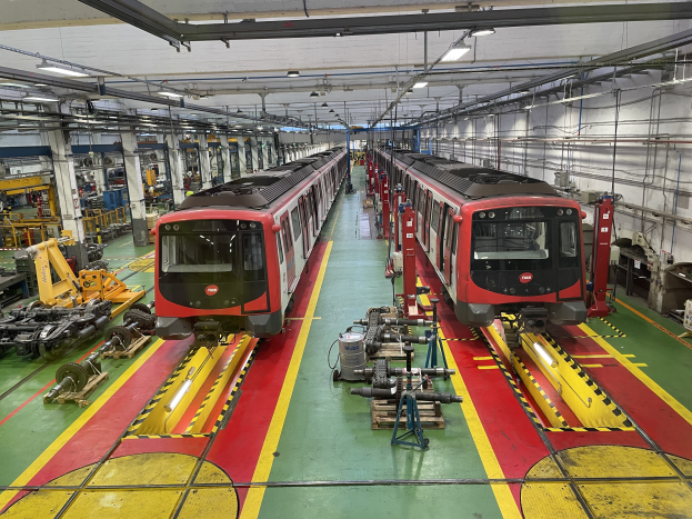 Zwei Züge auf den Schienen in einer Daimler Group-Fabrik, umgeben von Maschinen, Rohren und Deckenleuchten.