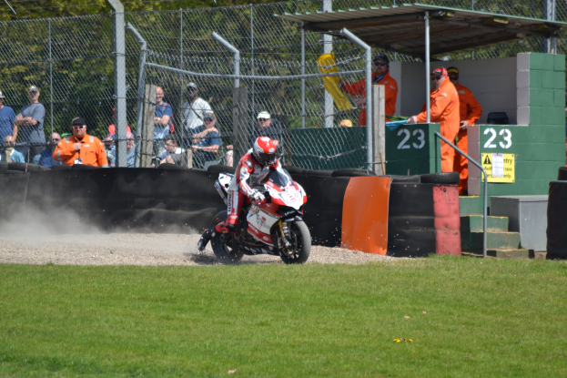Eine Person in Helm und Handschuhen auf einem Motorrad auf einem grasbewachsenen Feld, mit Zuschauern in Kappen und Schutzbrillen, einem eingezäunten Bereich, Treppen mit Geländern und einem nummerierten Schuppen im Hintergrund.