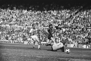 Schwarzes Foto von Fußballspielern im Einsatz auf einem Feld mit Zuschauern im Hintergrund und einem Bundesliga-Spielbanner von 1978-79.