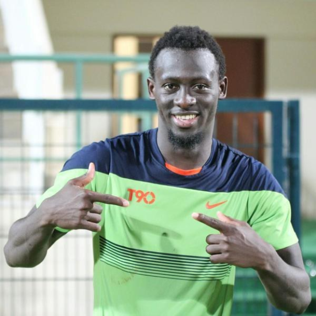 Ein Mann in einem grünen und blauen Hemd lächelt und zeigt auf die Kamera, mit einem unscharfen Geländer, Wand und Tür im Hintergrund.