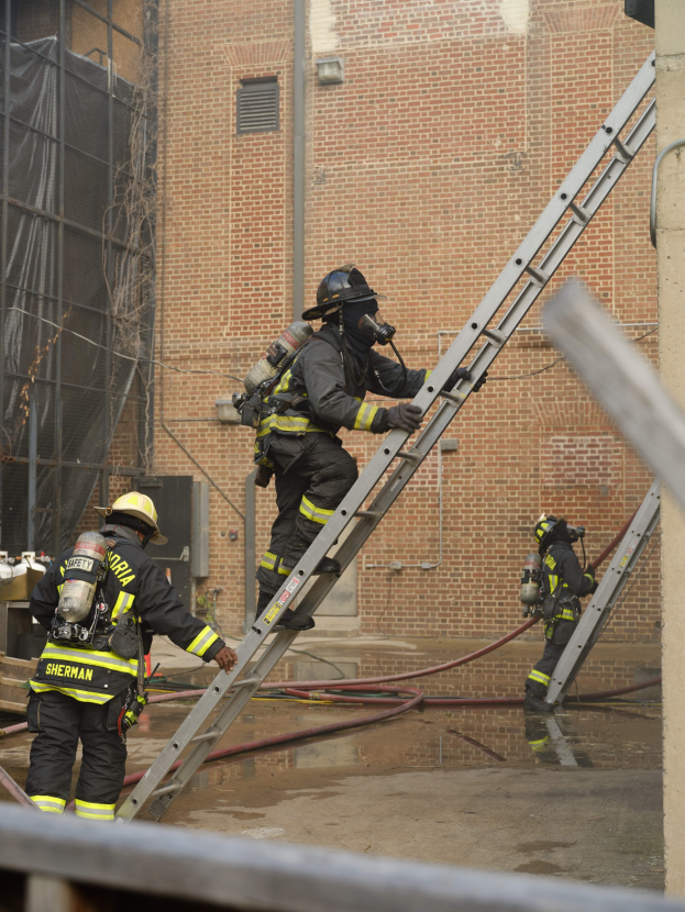 Feuerwehrleute in Helmen und Ausrüstung klettern an einer Leiter an einem Backsteingebäude mit Rohren auf dem Boden und einem Metallstab an der Basis hoch, während im Hintergrund ein weiteres Gebäude mit Fenstern und einem Netz zu sehen ist.