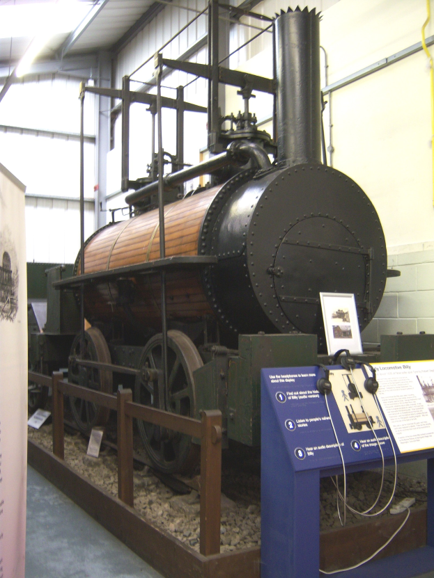 Alte Dampflokomotive in einem Museum mit Informationsschildern, Zaun und Deckenbeleuchtung ausgestellt.