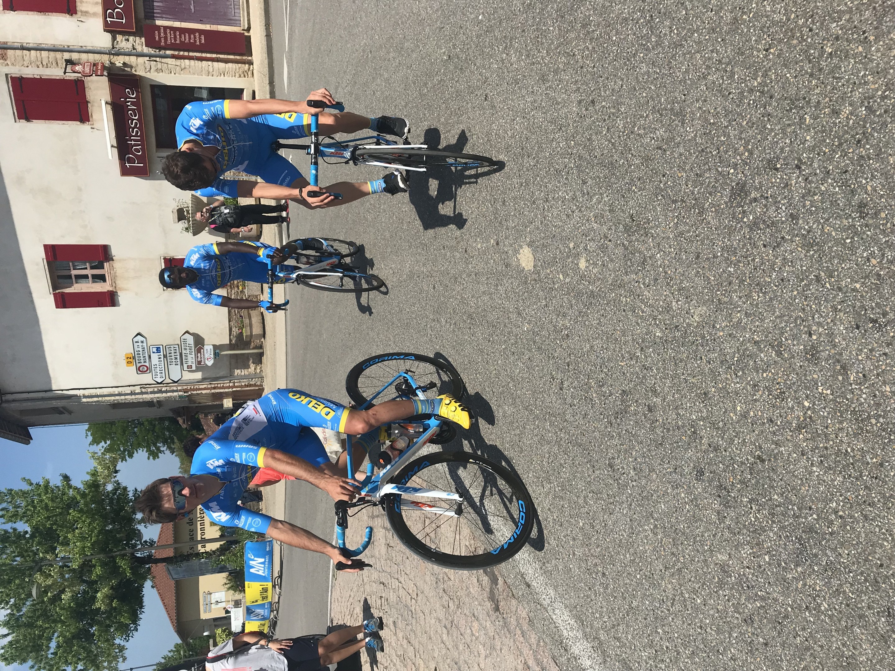 Eine Gruppe von Radfahrern fährt eine Straße entlang an einem Gebäude mit Fenstern und Texttafeln vorbei, mit Bäumen und einem klaren Himmel im Hintergrund und Zuschauern auf der Straße darunter.