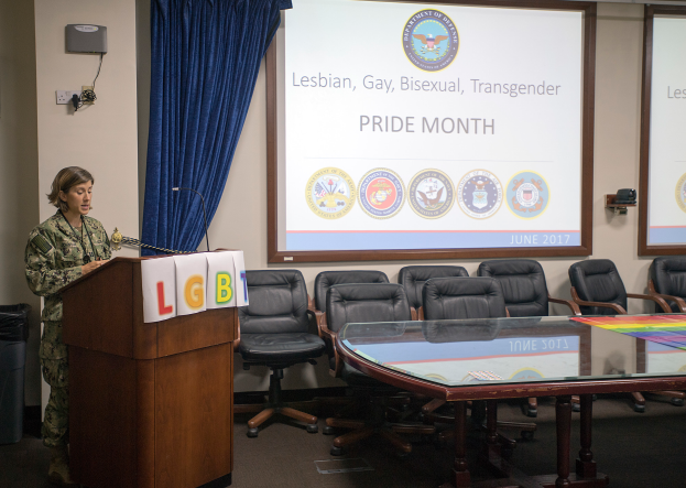 US Air Force Chief of Staff Gen. Kelly steht an einem Podium mit einem Mikrofon, während sie eine Gruppe vor einer Projektionswand anspricht, auf der "Pride Month" steht, mit Stühlen, einem Tisch mit Papieren, einem Vorhang, einem Lautsprecher, einem an der Wand montierten Gerät und Deckenleuchten im Hintergrund.