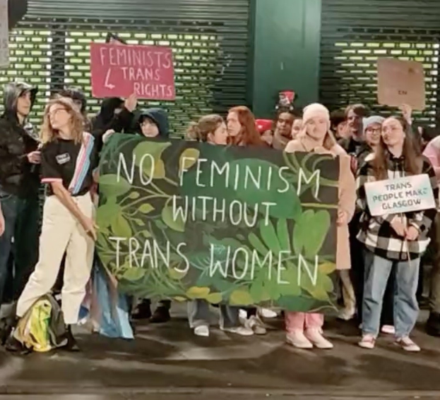 Eine Gruppe von Menschen hält ein Transparent mit der Aufschrift "Kein Feminismus ohne Trans-Frauen" und Schilder in einem öffentlichen Raum mit einer Wand und einem Rollladen im Hintergrund.