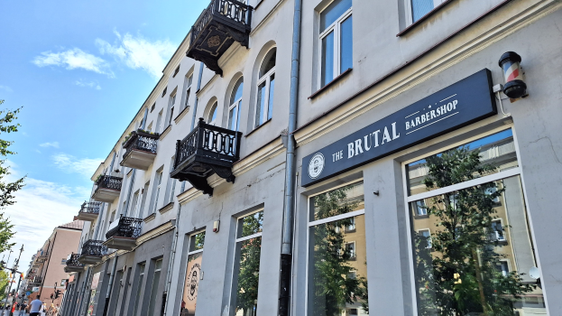Brutalistischer Barbershop in Berlin mit Betongebäuden, Fenstern, Geländern, Balkonen und Namensschildern, umgeben von Bäumen, Laternenmasten und Fußgängern unter einem bewölkten Himmel.