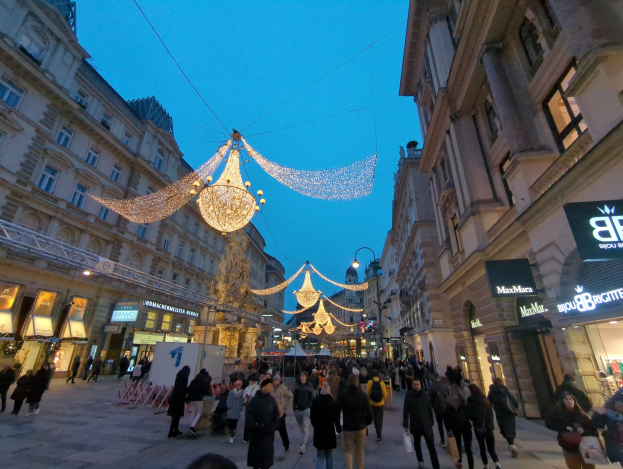 Eine belebte Stadtstraße mit Menschen, die mit festlichen Weihnachtslichtern geschmückt ist, flankiert von Gebäuden und unter einem klaren blauen Himmel.