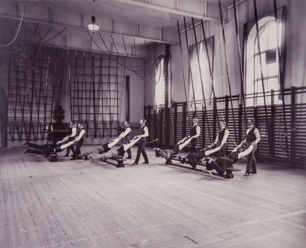 Schwarzes und weißes Bild einer Gruppe von Menschen, die Pilates-Übungen in einem Fitnessstudio mit einer Wand im Hintergrund, Fenstern auf der rechten Seite und Deckenlampen machen.