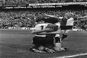 Drei Fussballspieler umarmen sich auf dem Boden nach einem Tor, mit jubelnder Menge und schwenkenden Fahnen in einem schwarz-weissen Stadion Setting.