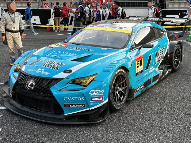 Blauer Lexus RC F GT3 Rennwagen auf einer Straße mit Zuschauern, einer Verkehrsinsel rechts und Hintergrundwerbung und Geländern.