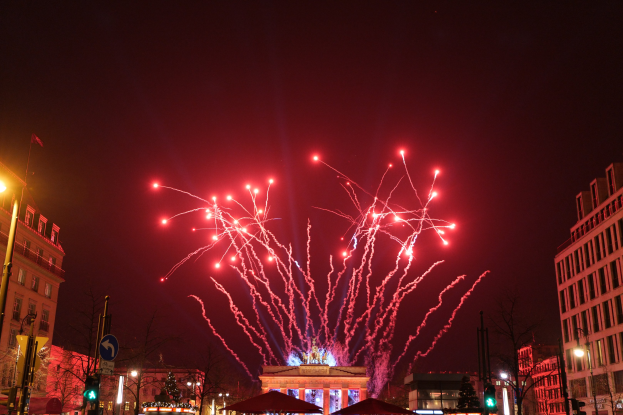 Eine belebte Stadtstraße an einem Neujahrsabend in Berlin mit Gebäuden, Bäumen, Laternen, Verkehrszeichen, Schildern, Zelten, Menschen und einem prächtigen Feuerwerk am Himmel.