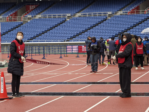 Gruppe von Menschen auf einer Bahn, die ein Band halten, mit einem Verkehrskegel links, Stühlen und Bannern im Hintergrund, was den Start eines Rennens anzeigt.