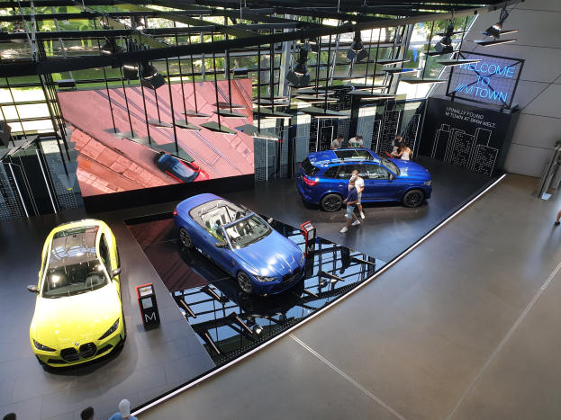 Eine Gruppe von Menschen, die im BMW Museum in München stehen, mit Autos auf der linken Seite und einem Schild auf der rechten Seite mit der Aufschrift "BMW Museum München", überblicken durch Glasfenster B├Ąume, Geb├Ąude und den Himmel.