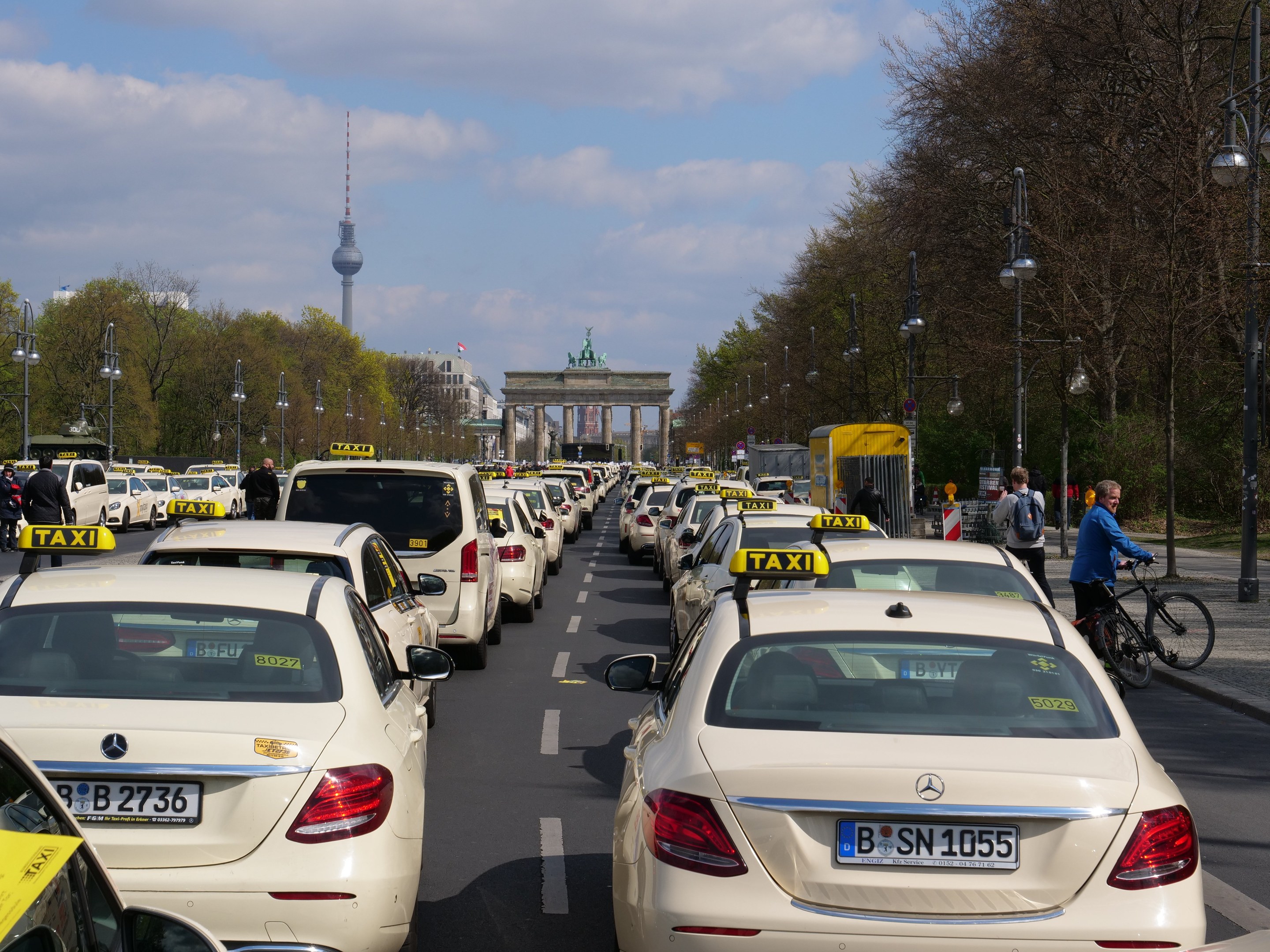 Eine lange Reihe von Taxis, die an einer belebten Straße in Berlin, Deutschland, geparkt sind, mit Fahrradfahrern und Fußgängern auf dem Gehweg, umgeben von Bäumen, Laternenmasten und Gebäuden, einschließlich eines Bogens und eines Turms unter einem bewölkten Himmel.