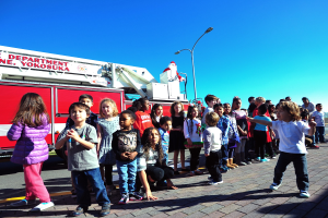 Eine Gruppe von Kindern vor einem Feuerwehrauto, das als Teil des Weihnachtsparades der Yokosuka Fire Department beschriftet ist, mit Laternenmasten, Bäumen und einem klaren blauen Himmel im Hintergrund.