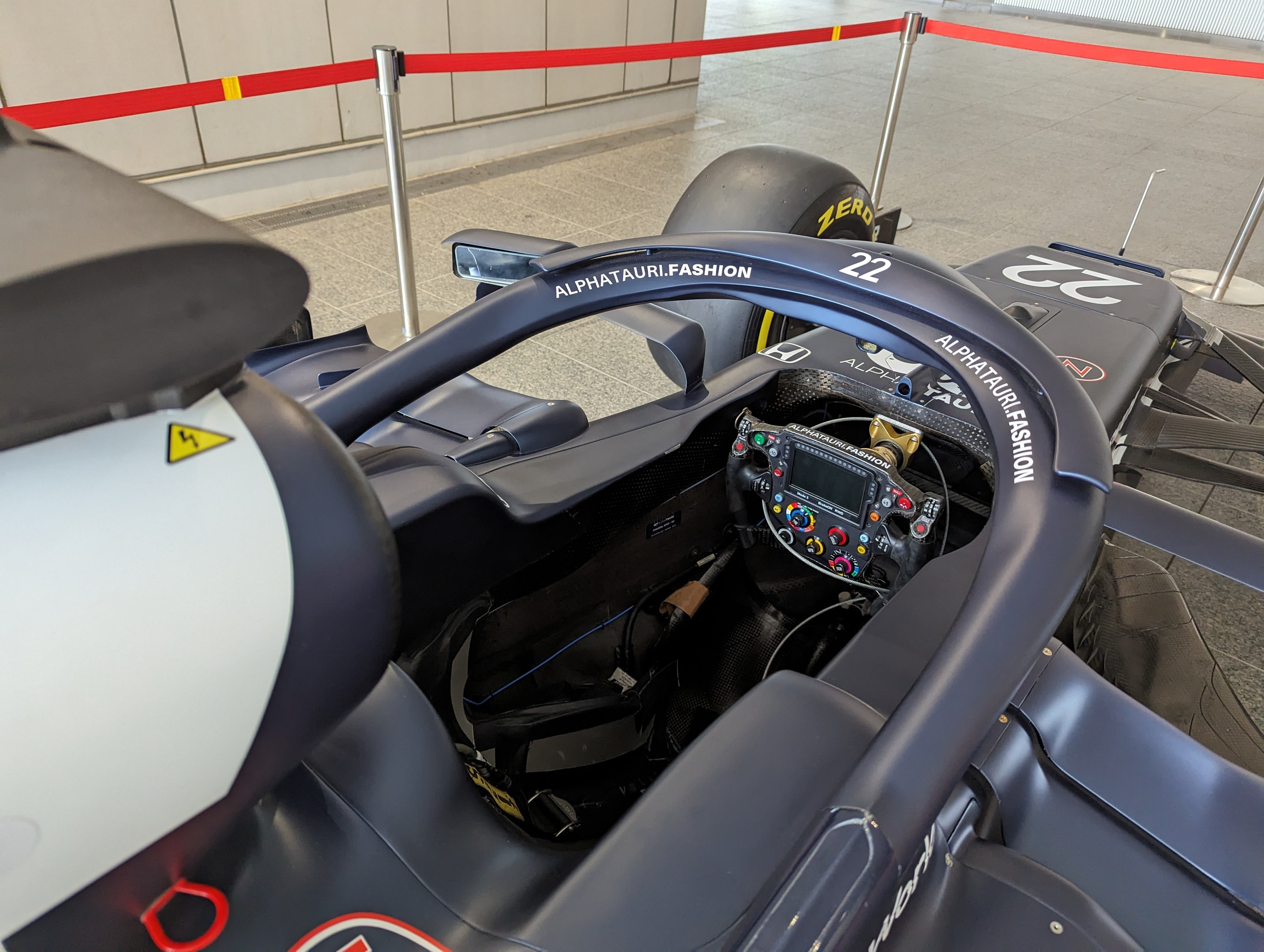 Cockpit eines Formel-E-Autos in einer Ausstellung mit einem Lenkrad im Vordergrund, einer Wand im Hintergrund und dekorativen Bändern um die Poles herum.