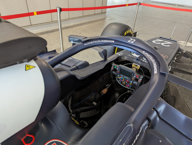 Cockpit eines Formel-E-Autos in einer Ausstellung mit einem Lenkrad im Vordergrund, einer Wand im Hintergrund und dekorativen Bändern um die Poles herum.