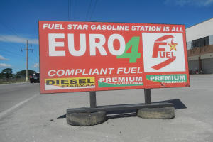 Eine Tankstelle mit einem 'Euro4 Fuel'-Schild im Vordergrund, Fahrzeuge auf der Straße, Strommasten mit Drähten, Bäume, ein Gebäude auf der rechten Seite und ein sichtbarer Himmel.