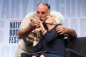 Ein Mann und eine Frau sitzen auf einem Sofa und umarmen sich, mit der Frau in einem blauen Kleid und einem weißen Banner mit der Aufschrift "National Book Festival" im Hintergrund.