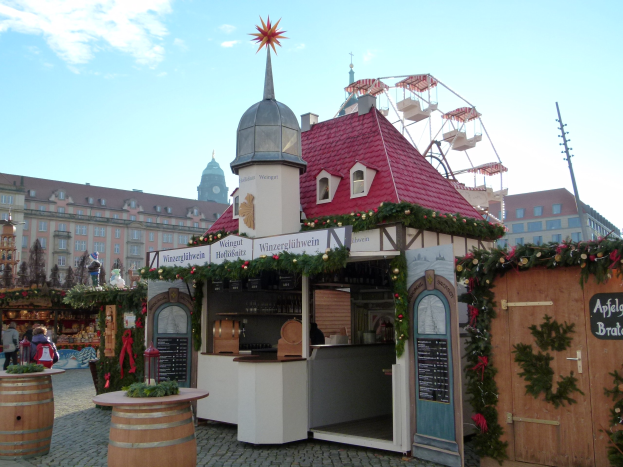 Kleines Gebäude mit rotem Dach und Riesenrad davor, umgeben von Festdekorationen und Menschen, mit Gebäuden, Bäumen und einem bewölkten Himmel im Hintergrund.