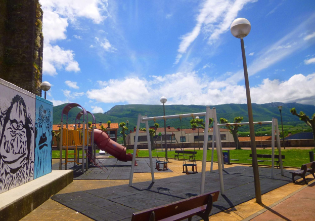 Ein Spielplatz mit Schaukeln, einer Rutsche, Bänken, Masten, Lichtern, einer bemalten Wand, Gras, Bäumen, Gebäuden, Hüllen und einem bewölktem Himmel im Hintergrund.
