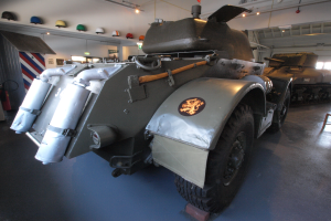 Ein gepanzertes Fahrzeug steht in einem Gebäude, umgeben von anderen Militärfahrzeugen, mit einem Feuerlöscher, einer Tafel und Helmen an der linken Wand und Beleuchtung von der Decke.