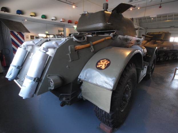 Ein gepanzertes Fahrzeug steht in einem Gebäude, umgeben von anderen Militärfahrzeugen, mit einem Feuerlöscher, einer Tafel und Helmen an der linken Wand und Beleuchtung von der Decke.