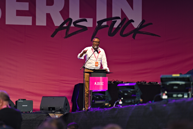 Ein Mann in einem weißen Hemd und braunen Hosen steht auf einer Bühne und hält ein Mikrofon in der Hand, mit einem pinken Banner hinter ihm, auf dem 'Berlin as Fuck' steht, und Lautsprechern auf der Bühne.