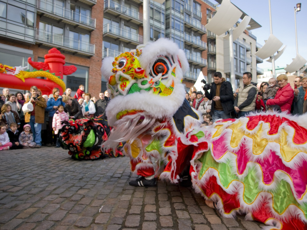 Vibrantes chinesisches Neujahrsfest in Amsterdam mit Löwen tanzen und einer Menge Schaulustiger, einige mit Kameras, vor einer Kulisse aus Gebäuden, Laternenmasten und einem klaren blauen Himmel.