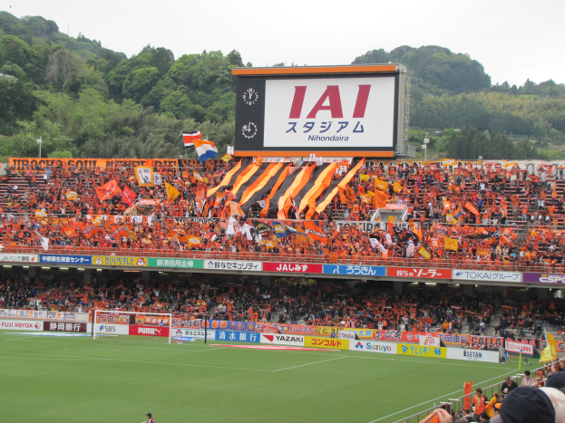 Ein Fussballspiel wird in einem Stadion mit einer grossen Zuschauermenge, grünem Rasen, einem Tor, Bannern, Fahnen, einem grossen Bildschirm, Bäumen und einem klaren blauen Himmel gespielt.