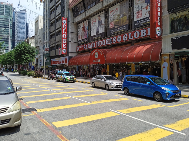 Eine belebte Stadtstraße mit Fahrzeugen, Fußgängern, Gebäuden, Schildern, Bannern, Schaufensterpuppen, Straßenmöblierung, Grünflächen und einem bewölkten Himmel.