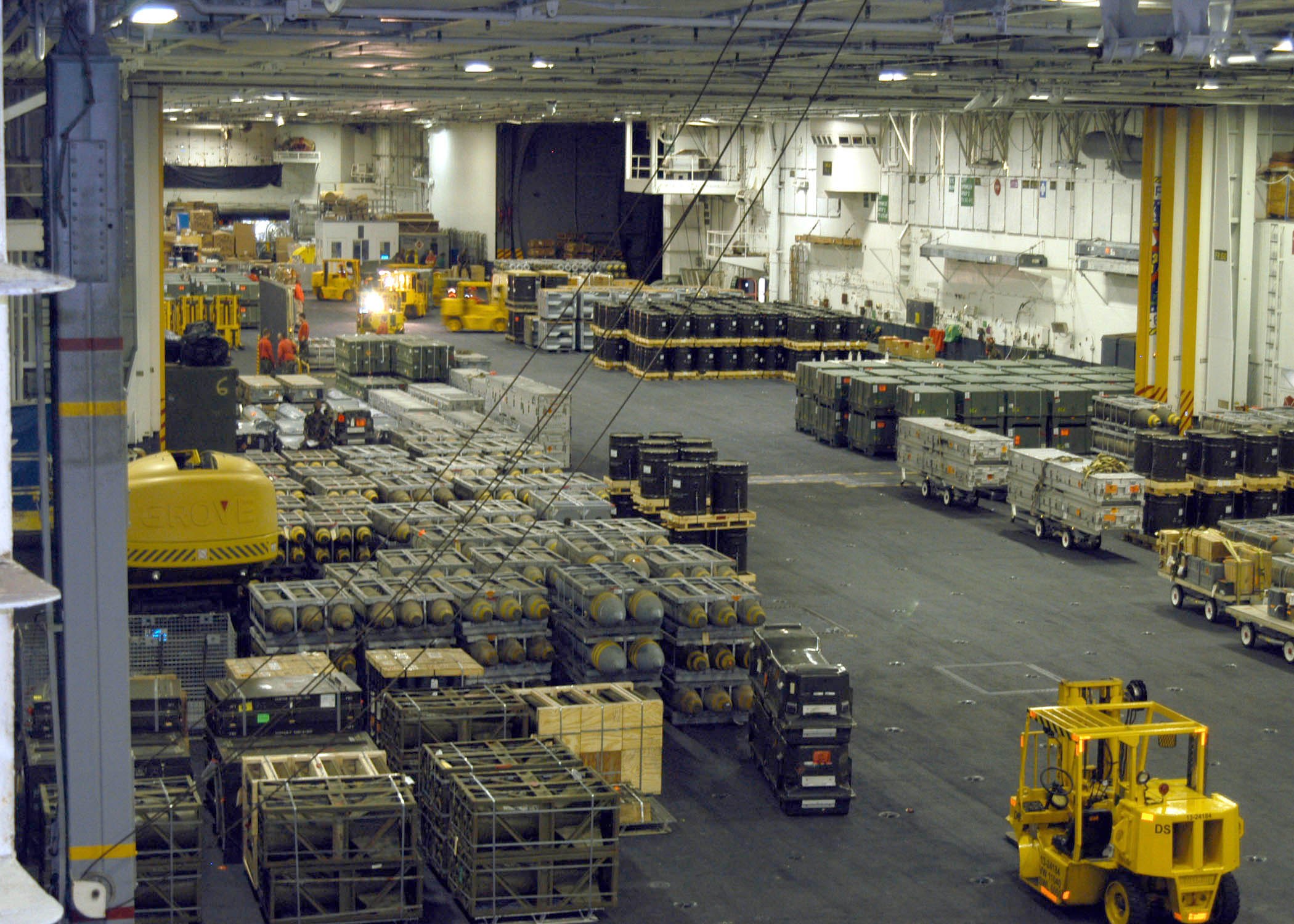 Großer Lagerinnenraum mit mehreren Lastwagen, Containern, Menschen, Deckenbeleuchtung, Metallstangen und einem sichtbaren Rad.