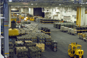 Großer Lagerinnenraum mit mehreren Lastwagen, Containern, Menschen, Deckenbeleuchtung, Metallstangen und einem sichtbaren Rad.