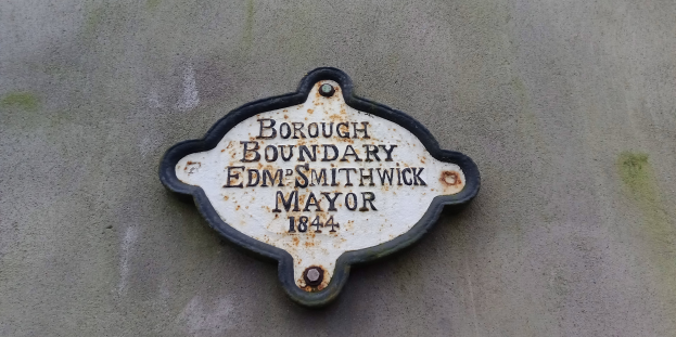 Metallschild an einem Gebäude mit schwarzem Hintergrund und weißer fettgedruckter Schrift, die "Borough Boundary Edm Smithwick Mayor" liest.