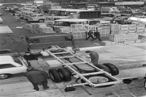 Schwarze und weiße Fotografie von Männern, die an einem Lkw in einer Fabrik arbeiten, mit Fahrzeugen, Menschen und Kisten im Hintergrund.