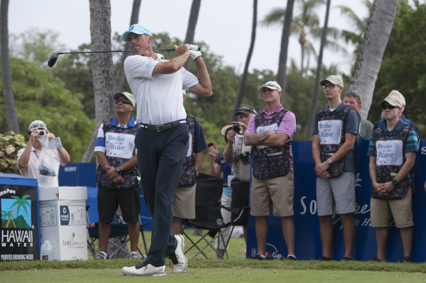 Ein Mann in einem weißen T-Shirt, schwarzen Hosen und einer blauen Mütze schwingt einen Golfschläger auf einem grünen Feld, umgeben von Zuschauern und Kameras unter einem klaren blauen Himmel.
