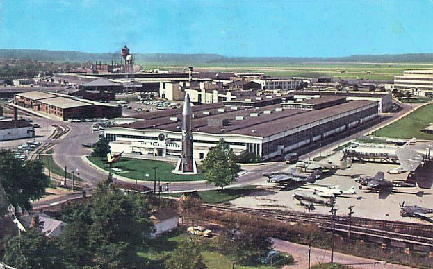 Altes Schwarz-Weiß-Foto eines Flughafens mit parkenden Flugzeugen, Gebäuden, Bäumen, Straßen, Fahrzeugen, Strommasten, Gras, Bahnschienen, Türmen, fernen Hügeln und einem klaren blauen Himmel; Text erscheint unten.