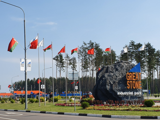 Großer Fels mit der Inschrift "Großer Stein Industriepark" in einer parkähnlichen Anlage mit Gras, Blumen, Bäumen, Laternen mit Flaggen, Gebäuden und einem blauen Himmel.