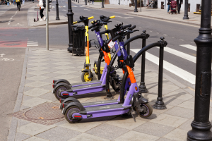 Eine Reihe von Elektro-Scootern, die auf der Stra\ssenseite geparkt sind, mit Menschen, die auf dem Gehweg gehen und Fahrr"ader fahren, ein M"ullbeh"alter im Vordergrund und Geb"aude, B"ume und Pflanzen im Hintergrund.
