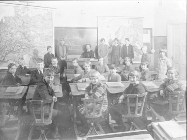 Schwarzes und weißes Foto eines Klassenzimmers mit Kindern auf Stühlen sitzend und stehend, Bücher auf Tischen, Landkarten an der Wand und eine Tür auf der rechten Seite.