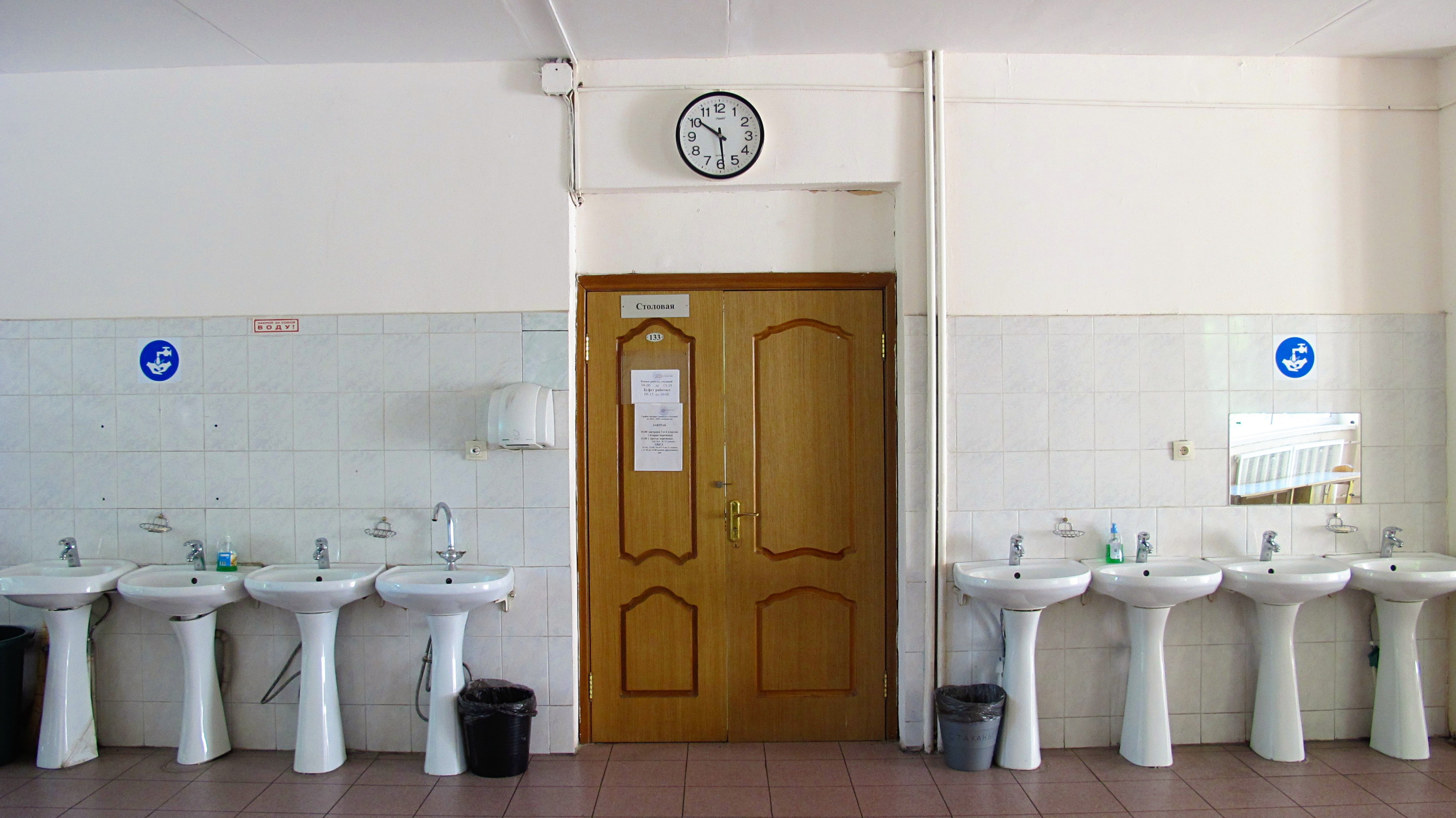 Reihe von Waschbecken mit Wasserhähnen, Mülltonnen auf dem Boden, eine Tür im Hintergrund, eine Uhr an der Wand und ein Spiegel auf der rechten Seite in einem Badezimmer.