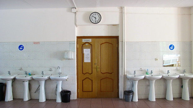 Reihe von Waschbecken mit Wasserhähnen, Mülltonnen auf dem Boden, eine Tür im Hintergrund, eine Uhr an der Wand und ein Spiegel auf der rechten Seite in einem Badezimmer.