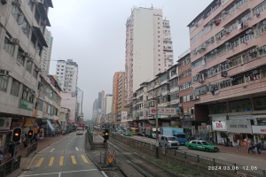 Eine belebte Stadtstraße mit hohen Gebäuden, fahrenden Fahrzeugen, Fußgängern, Ampeln, Schildern, Geländern und einem Himmel im Hintergrund.
