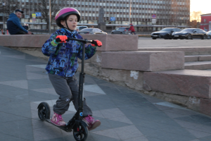 Ein junger Junge fährt mit einem Helm und Handschuhen auf einem Gehweg mit einem E-Scooter, mit verschiedenen städtischen Elementen und einem klaren blauen Himmel im Hintergrund.