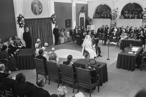 Ein Schwarz-Weiß-Foto einer Hochzeitszeremonie in einem großen Raum mit Gästen auf Stühlen und Stehplätzen, Tischen mit Papieren und Mikrofonen, einer dekorierten Wand mit Foto-Rahmen und Blumenbouquets und einem Podium mit Mikrofonständer.
